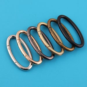 May include: A selection of oval metal clasps in different metallic finishes, such as silver, bronze, and black, set against a solid blue backdrop. These clasps are suitable for bags, jewellery, or other craft projects.