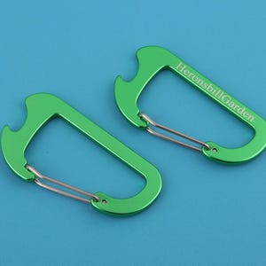 以下が含まれることがあります： 緑色のカラビナクリップ2つ。ボトルオープナー機能とシルバーのスプリングゲート付き。1つのクリップには「HeronsbillGarden」の文字がプリントされています。クリップは青色の背景にあります。