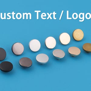 May include: A collection of round metal buttons in various colors, including black, silver, gold, and bronze, arranged on a blue background. The text "Custom Text / Logo" is displayed at the top. These buttons are likely for clothing or craft projects.