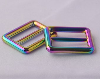 Op de afbeelding: Twee regenboogkleurige metalen schuifgespen. De gespen zijn rechthoekig en hebben een gladde, glanzende afwerking. De kleur is een regenboogeffect met tinten van paars, blauw, groen, geel en oranje.