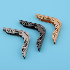 May include: Three decorative metal corner protectors with an ornate design. The corner protectors are in gold, silver, and black.