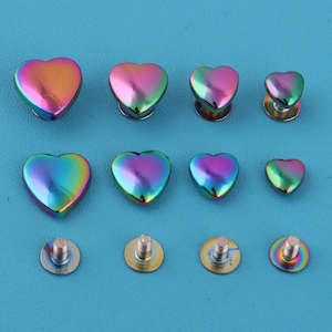 Könnte beinhalten: Eine Sammlung herzförmiger Metallnieten in verschiedenen Größen, angeordnet auf einem blauen Hintergrund. Die Nieten haben eine lebendige, schillernde Oberfläche mit Farben von Rosa und Lila bis Grün und Gold. Die Nieten sind zur Befestigung mit einer Schraube konzipiert.