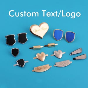 May include: An assortment of metal embellishments in various shapes, including hearts, shields, and rectangular bars. Some pieces are gold-toned, others are black or blue. The text "Custom Text/Logo" is at the top.
