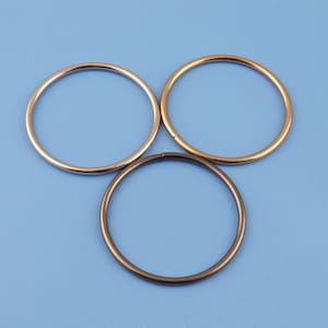 May include: Three metal rings in gold, bronze, and brass colors are arranged against a light blue background. The rings are circular and appear to be made of a smooth, shiny metal. The rings are likely used for crafting or jewelry making.