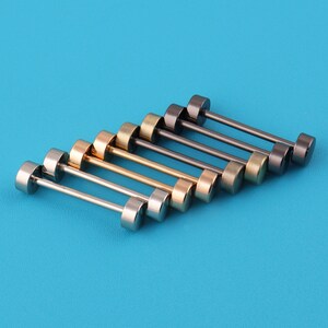 May include: An assortment of barbell-style stud earrings in various metallic finishes, including silver, gold, and gunmetal. The earrings are arranged diagonally against a solid blue background, showcasing their cylindrical design.