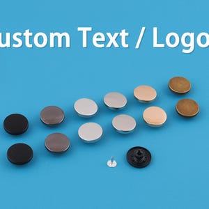 May include: A collection of metal snap fasteners in various colors, including black, silver, gold, and bronze, arranged on a blue background. The text "Custom Text / Logo" is displayed at the top. Includes a snap fastener with a pin.