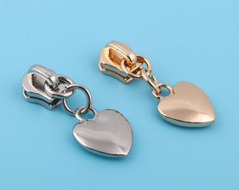 May include: Two metal zipper pulls with heart-shaped charms. One pull is silver-toned, the other is gold-toned. Both have a ring connecting the pull to the heart charm. The background is a solid light blue.