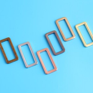 Könnte beinhalten: Rechteckige Metallringe in verschiedenen Farben, darunter Bronze, Silber, Kupfer und Gold, auf blauem Hintergrund. Diese Hardware-Komponenten werden wahrscheinlich für Bastel- oder Taschenprojekte verwendet.