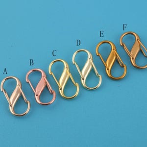 May include: Six metal S-shaped clasps in various finishes: rose gold, gold, and silver. The clasps are arranged on a blue background. Each clasp has a unique color and sheen, suitable for jewelry making or crafting.