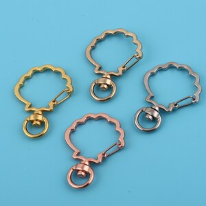 Könnte beinhalten: Vier Schlüsselanhänger aus Metall in Form einer Muschel mit einem Drehverschluss. Die Schlüsselanhänger sind golden, silber, roségold und schwarz.