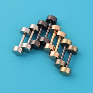 May include: An assortment of dumbbell-shaped stud earrings in various metallic finishes, including silver, black, and gold. The earrings are arranged on a bright blue surface, showcasing their cylindrical design and polished appearance.