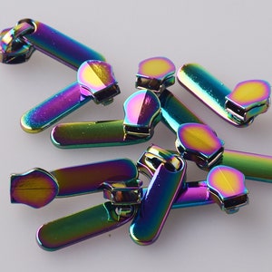 Puede incluir: Un grupo de tiradores de cremallera iridiscentes en varios colores arcoíris. Los tiradores están hechos de metal y tienen un acabado liso y brillante.