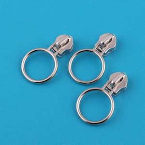 Op de afbeelding: Drie zilverkleurige ritstrekkers met een ringvormige handgreep. De trekkers zijn gemaakt van metaal en hebben een glanzende afwerking. De achtergrond is effen helderblauw. Deze ritstrekkers zijn waarschijnlijk voor kleding of tassen.