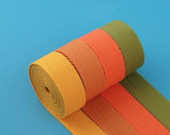 May include: A roll of colorful woven fabric in shades of yellow, orange, and olive green. The fabric is tightly woven with a textured appearance. The roll is positioned diagonally against a solid blue background, showcasing the different colors.