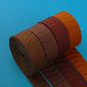 Puede incluir: Un rollo de tela tejida en tonos marrón, burdeos y naranja sobre un fondo azul. La tela parece ser un material resistente, posiblemente lona o cincha, adecuado para diversos usos artesanales o industriales.