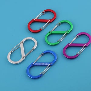 May include: Five colorful S-biner carabiners in red, green, purple, blue, and silver. The carabiners are made of metal and have a spring-loaded gate. The blue carabiner has the text "HeronsbillGarden".