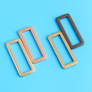 May include: Four rectangular metal rings in gold, copper, and bronze tones are arranged on a bright blue background. The rings are open in the centre, and the metal has a smooth, polished finish. These rings could be used for various craft projects.