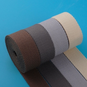 Può includere: Un rotolo di fettuccia tessuta nei toni del marrone, grigio e beige su sfondo blu. La fettuccia è fittamente tessuta con una superficie strutturata e i colori sono disposti a strisce parallele.