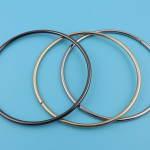 May include: Three metal hoop necklaces in black, gold, and silver, arranged on a light blue background. The necklaces are circular with a simple, minimalist design, and appear to be made of thin metal tubing.