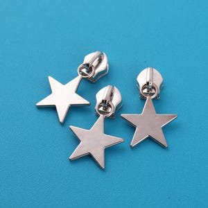 Op de afbeelding: Zilverkleurige ritstrekkers met stervormige bedels. De ritstrekkers zijn van metaal en hebben een glanzende afwerking. De sterbedels zijn plat en hebben vijf punten. De achtergrond is effen helderblauw.