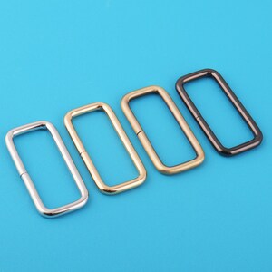 Puede incluir: Cuatro anillos metálicos rectangulares en plata, oro y negro, dispuestos diagonalmente sobre un fondo azul brillante. Los anillos parecen ser componentes de hardware, posiblemente para bolsos u otros proyectos de artesanía.