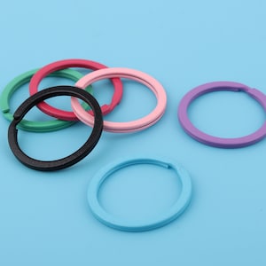 May include: Five colorful metal key rings in pink, green, black, and blue. The key rings are all different sizes and are arranged in a circle on a blue background.