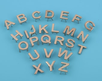 May include: A set of silver metal alphabet letter charms with a pointed back for attaching to fabric or other materials.