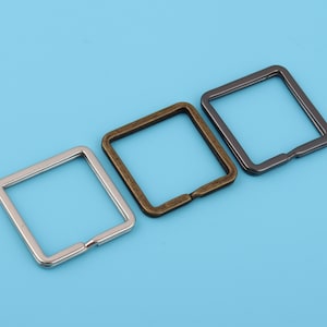 May include: Three square metal rings in different finishes: silver, bronze, and black. The rings are all the same size and shape, and they are arranged in a row from left to right.