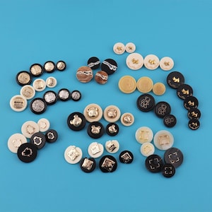 May include: A collection of various decorative buttons in different sizes and designs, including gold, silver, and black accents. Some buttons feature bear, dog, and geometric shapes. The buttons are arranged on a bright blue background.