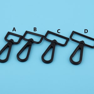 Puede incluir: Cuatro ganchos giratorios de metal negro etiquetados como A, B, C y D. Los ganchos son todos del mismo tamaño y forma, con una abertura rectangular en la parte superior y un gancho curvo en la parte inferior.