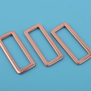 May include: Three rose gold metal rectangular belt buckle slides. The buckles are arranged in a row, with the largest buckle on the right and the smallest buckle on the left.