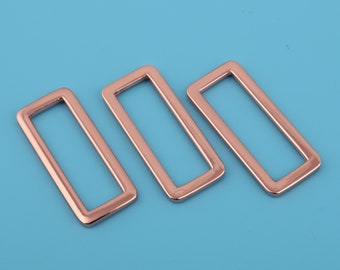 May include: Three rose gold metal rectangular belt buckle slides. The buckles are arranged in a row, with the largest buckle on the right and the smallest buckle on the left.