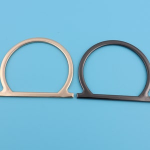 May include: Two metal bag handles, one silver and one black, are shown against a light blue background. The handles are shaped like an upside-down U.