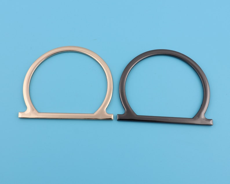 May include: Two metal bag handles, one silver and one black, are shown against a light blue background. The handles are shaped like an upside-down U.