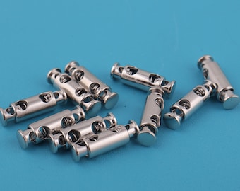 May include: A collection of shiny silver cord locks, also known as barrel or toggle cord locks, are scattered on a blue surface. These small, cylindrical fasteners have a hole for cord insertion and are commonly used in apparel and accessories.