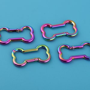Op de afbeelding: Vier iriserende karabijnhaakclips in de vorm van een hondenbot. De clips zijn gemaakt van metaal en hebben een regenboogafwerking, met kleuren die verschuiven van roze naar blauw en groen. Ze zijn gerangschikt op een effen blauwe achtergrond.