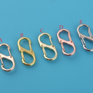 Könnte beinhalten: Fünf S-förmige Metallverschlüsse in verschiedenen Ausführungen: Roségold, Gold und Silber. Die Verschlüsse werden vor einem einfarbigen blauen Hintergrund dargestellt. Diese Verschlüsse werden häufig für die Schmuckherstellung, Schlüsselanhänger und andere Bastelprojekte verwendet.