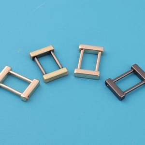 May include: Four metal rectangle-shaped hardware pieces in gold, bronze, and gunmetal gray. Each piece has a rectangular bar on each end, connected by two parallel bars. The hardware pieces are arranged on a blue surface.