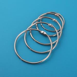 Puede incluir: Un conjunto de anillas de encuadernación de metal plateado, cada una con una abertura articulada, dispuestas sobre un fondo azul liso. Las anillas son circulares y parecen estar hechas de un metal brillante y duradero.