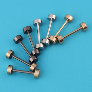 May include: An assortment of barbell stud earrings in various metallic finishes, including gold, silver, and dark gray. The earrings are arranged on a bright blue background, showcasing their cylindrical posts and circular ends. These earrings are suitable for everyday wear.