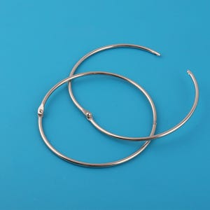 Puede incluir: Dos anillas plateadas, ligeramente abiertas, sobre un fondo azul liso. Las anillas son circulares con un mecanismo de apertura con bisagras. El metal tiene una superficie brillante y reflectante.