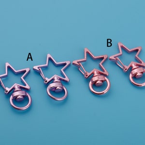 May include: Three star-shaped keychains with swiveling rings. The keychain on the left is a light purple colour, the middle keychain is a darker purple colour, and the keychain on the right is a rose gold colour.