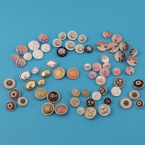 Puede incluir: Una colección de botones decorativos variados en diferentes tamaños y diseños. Los botones presentan una gama de colores, incluyendo dorado, plateado, rosa y negro, con patrones florales y geométricos. Algunos botones tienen apariencia de joya.