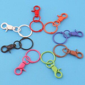 May include: A selection of brightly coloured metal keyrings with lobster claw clasps and split rings. The keyrings are in various colours, including red, orange, purple, green, pink, brown, black, and white, against a blue backdrop.