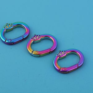 May include: Three rainbow-colored oval carabiner clips with a crown design on a blue background. The clips have a metallic finish and a spring-loaded gate for secure closure. These decorative clips are suitable for various crafting and accessory projects.