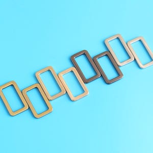 Op de afbeelding: Rechthoekige metalen ringen in goud, brons en zilver op een helderblauwe achtergrond. Deze hardware-componenten worden waarschijnlijk gebruikt voor het maken van tassen of andere knutselprojecten. De ringen zijn gelijkmatig verdeeld en hebben een strak, modern ontwerp.