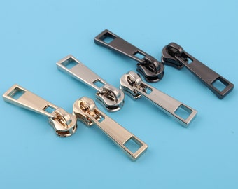 May include: Three zipper pulls in different finishes: gold, silver, and black. Each pull has a rectangular shape with a small hole at the top.