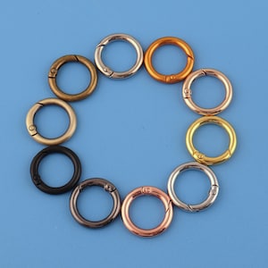Pode incluir: Uma coleção de dez fechos de mola redondos de metal em várias cores, incluindo dourado, prateado, bronze e preto, dispostos em círculo sobre um fundo azul. Estes fechos são usados para prender itens.