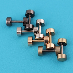 May include: An assortment of dumbbell-shaped stud earrings in various metallic finishes, including gunmetal, silver, gold, and bronze. The earrings are arranged on a vibrant blue background, showcasing their simple yet stylish design. These earrings are a unique accessory.