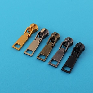 Op de afbeelding: Vijf metalen ritstrekkers in goud, brons en zwart op een blauwe achtergrond. Elke trekker heeft een rechthoekige basis met een vierkante opening en een gebogen bovenkant met een lus.